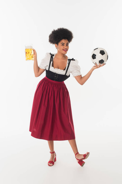 african american oktoberfest waitress with mug of foamy beer playing football on white, full length - Fotografie, Obrázek