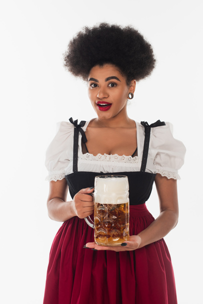 здивована афроамериканська баварська офіціантка в костюмі баварського мистецтва з кубком пікантного пива на білому - Фото, зображення