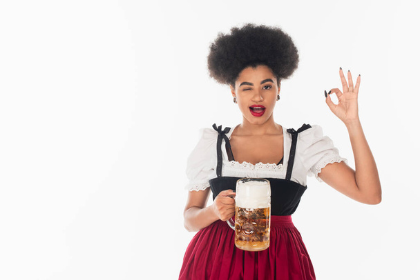 serveuse afro-américaine oktoberfest avec tasse de clin d'oeil à la bière artisanale et montrant un signe acceptable sur blanc - Photo, image