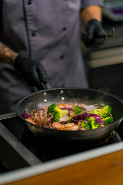 close-up van een chef-kok in een professionele keuken bereiden van zeevruchten saute mengen ingrediënten in pan gooien ze in de lucht - Foto, afbeelding