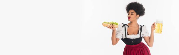 zadowolona afrykańska kelnerka bawarska z kubkiem i hot dogiem na białym, oktoberfest, baner - Zdjęcie, obraz
