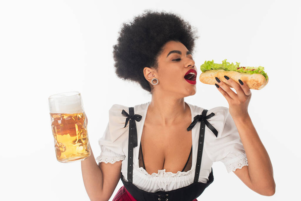 Африканская американская баварская официантка с кружкой пива, поедающая вкусный хот-дог на белом, октоберфест - Фото, изображение