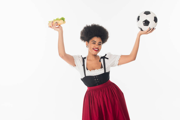izgatott afro-amerikai bajor pincérnő focilabdával és hot dog emelt kézzel fehér - Fotó, kép