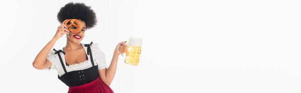 gelukkig Afrikaans amerikaans bavariaans serveerster met bier mok obscuring gezicht met pretzel op wit, banner - Foto, afbeelding