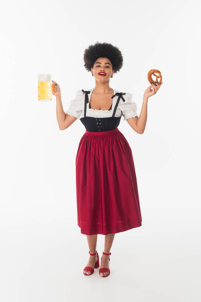 счастливая африканская американская баварская официантка в костюме Октоберфеста с кружкой пива и кренделем на белом - Фото, изображение