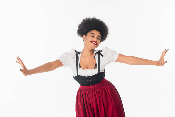 joyeuse serveuse afro-américaine en costume bavarois traditionnel avec les mains tendues sur blanc - Photo, image