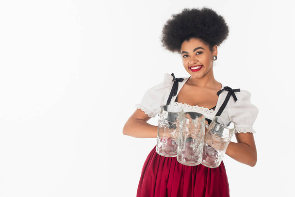 charming african american bavarian waitress in oktoberfest outfit holding empty beer mags on white - Foto, Imagem
