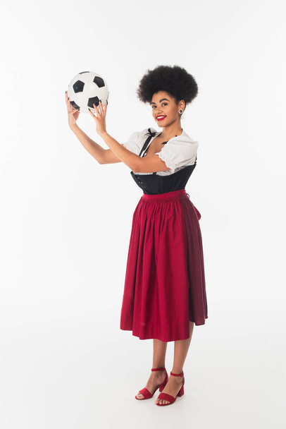 smiling african american bavarian waitress in authentic dirndl dress holding soccer ball on white - Foto, immagini