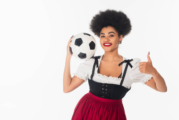 radostná afroamerická servírka oktoberfestu v dirndl držící fotbalový míček a ukazující jako na bílém - Fotografie, Obrázek