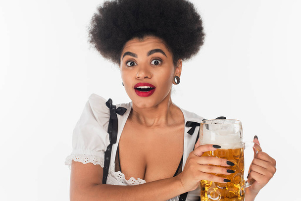 surpris serveuse afro-américaine dans dirndl bavaroise traditionnelle tenant tasse de bière sur blanc - Photo, image
