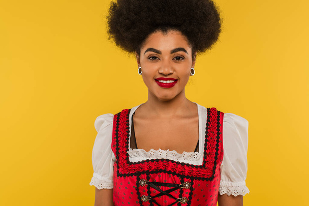 aantrekkelijke Afrikaans-Amerikaanse bavariaanse serveerster in traditioneel kostuum op zoek naar camera op geel - Foto, afbeelding