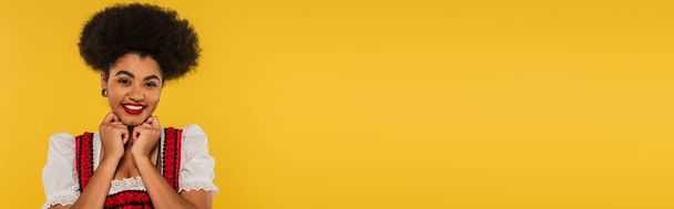 возбужденная африканская американская баварская официантка в традиционной одежде для фестиваля Октоберфест на желтом, баннер - Фото, изображение