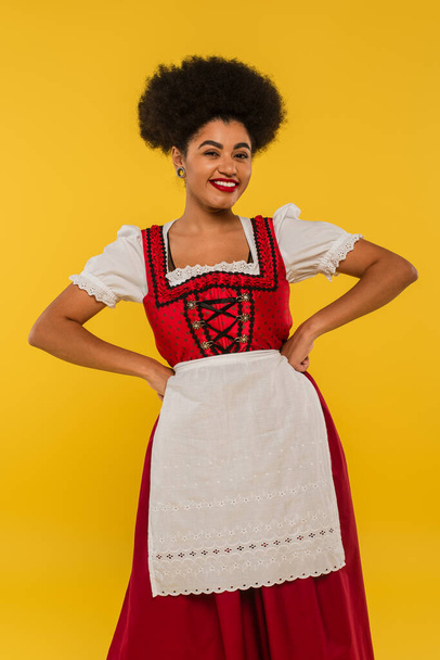 cheerful african american bavarian waitress in dirndl holding hands on waist on yellow, oktoberfest - Foto, Bild
