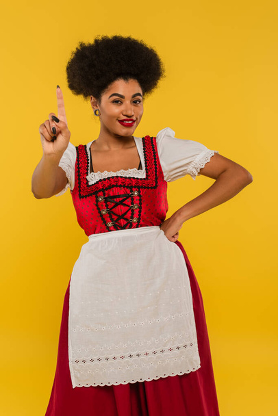 sonriente africana americana camarera bavariana con la mano en la cadera mostrando gesto de idea en amarillo - Foto, Imagen