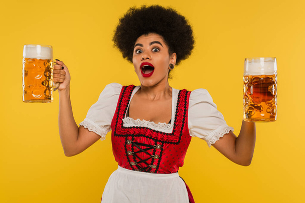 emocionado camarera bavariana afroamericana con la boca abierta de pie con revistas de cerveza completa en amarillo - Foto, Imagen