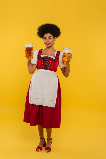african american waitress in oktoberfest dirndl holding beer mugs and smiling at camera on yellow - Photo, Image