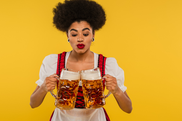 joven africano americano oktoberfest camarera en bavariana traje tazas tintineo con cerveza en amarillo - Foto, Imagen