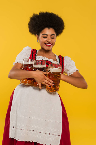 positivo africano americano bavarian camarera en dirndl holding completo cerveza tazas y sonriendo en amarillo - Foto, imagen