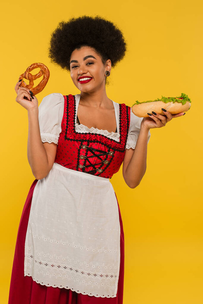 pretty african american oktoberfest waitress with hotdog and pretzel smiling at camera on yellow - Foto, afbeelding