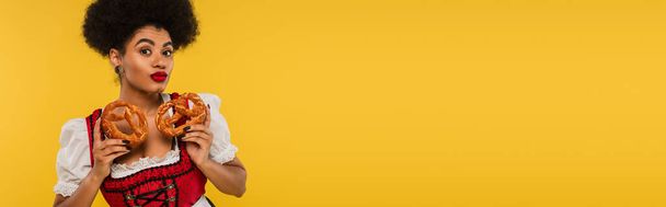 jolie serveuse afro-américaine oktoberfest avec de savoureux bretzels regardant caméra sur jaune, bannière - Photo, image