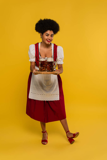charming african american oktoberfest waitress with beer mugs on wooden tray smiling on yellow - Foto, immagini