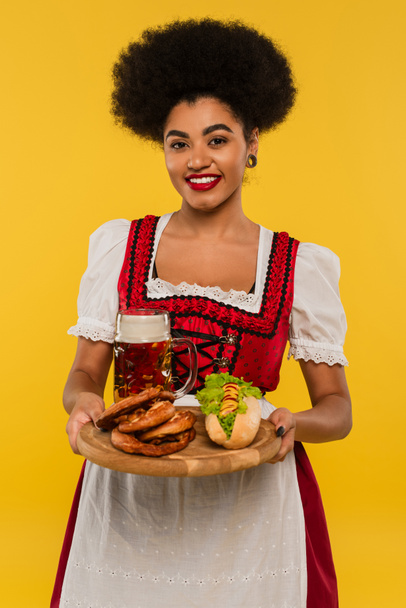 joyful african american oktoberfest waitress holding wooden tray with pretzels and hot dog on yellow - Fotó, kép