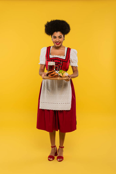 афроамериканська офіціантка, що тримає дерев'яний піднос з пивом і смачно ласощі на жовтому - Фото, зображення