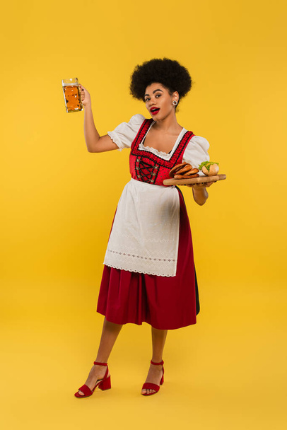 Afrikaans Amerikaans bavariaans serveerster in dirndl poseren met bier en eten op houten dienblad op geel - Foto, afbeelding