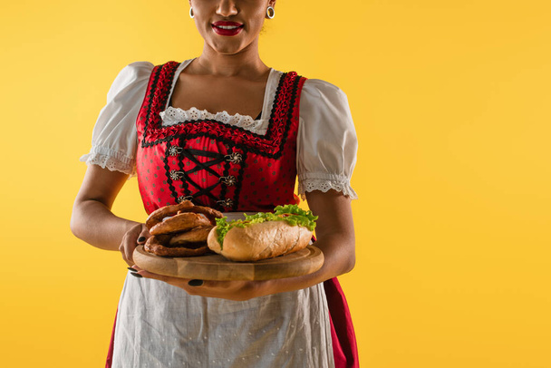 cropped african american bavarian waitress with pretzels and hot dog on wooden tray on yellow - Photo, Image