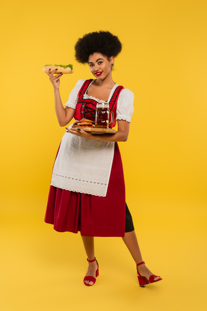 alegre afroamericana oktoberfest camarera con cerveza y deliciosa golosina en bandeja de madera en amarillo - Foto, Imagen