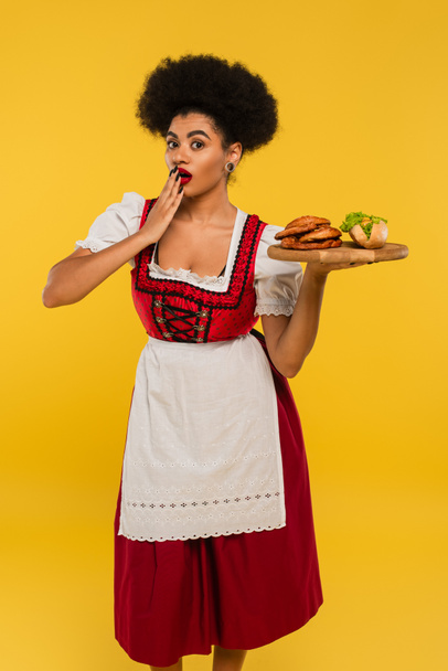 шокована афроамериканська баварська офіціантка тримає дерев'яний піднос з кренделями та хот-догом на жовтому - Фото, зображення