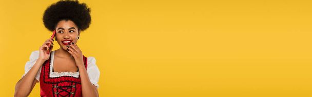 счастливая африканская американская баварская официантка в аутентичной одежде разговаривает по смартфону на желтом, баннер - Фото, изображение