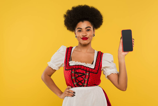 afro-amerikai oktoberfest pincérnő dirndl mutatja okostelefon üres képernyő sárga - Fotó, kép
