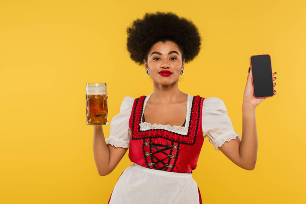 camarera bavariana afroamericana con taza de cerveza y teléfono inteligente con pantalla en blanco en amarillo - Foto, Imagen