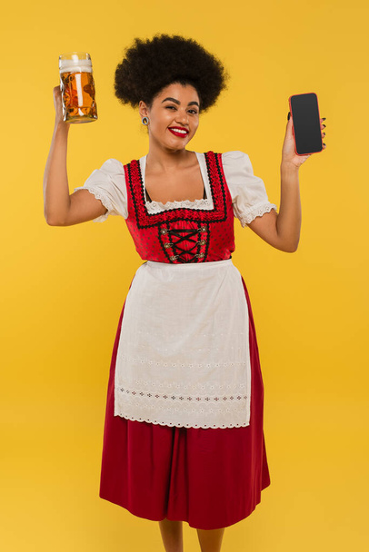 Mutlu Afro-Amerikan Bavyera garsonu bira bardağı ve cep telefonuyla sarı ekranlı. - Fotoğraf, Görsel
