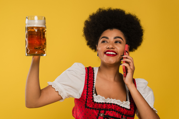 excited african american oktoberfest waitress with beer mug talking on smartphone on yellow - Фото, зображення