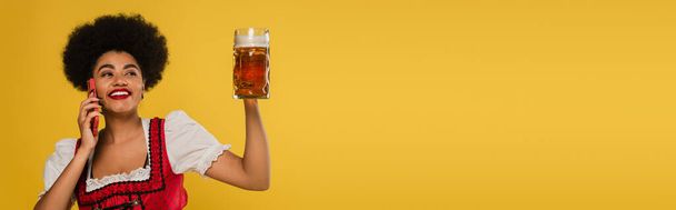 счастливая африканская американская баварская официантка с кружкой пива разговаривая по смартфону на желтом, баннер - Фото, изображение