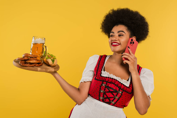afrikanisch-amerikanische bayrische Kellnerin mit Bier und Snacks auf Tablett im Gespräch auf dem Smartphone auf gelb - Foto, Bild
