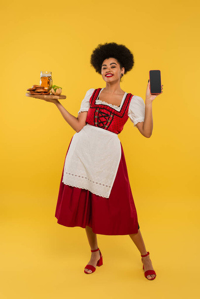 africká americká bavorská servírka s pivem a jídlem zobrazující prázdnou obrazovku smartphonu na žluté - Fotografie, Obrázek