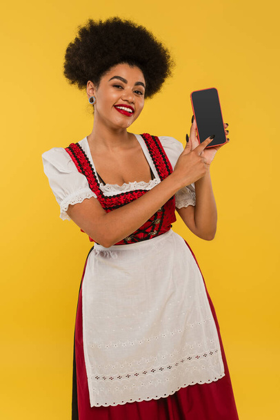 camarera bavariana afroamericana feliz en dirndl apuntando a la pantalla en blanco del teléfono inteligente en amarillo - Foto, imagen