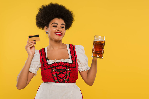 alegre africano americano bavarian camarera en dirndl traje con cerveza taza y tarjeta de crédito en amarillo - Foto, imagen