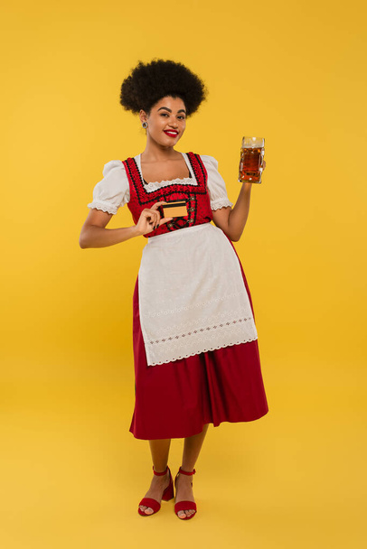 happy african american bavarian waitress in dirndl dress with beer mug and credit card on yellow - Фото, зображення