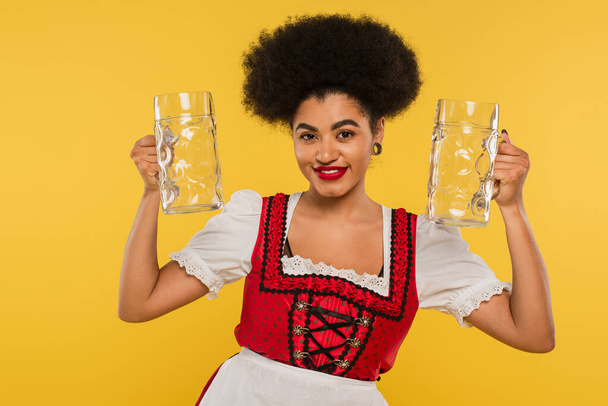 pleased african american oktoberfest waitress in dirndl costume holding empty beer mugs on yellow - Foto, Bild