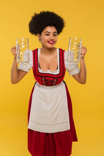 happy african american oktoberfest waitress in dirndl dress holding empty beer mugs on yellow - Photo, Image