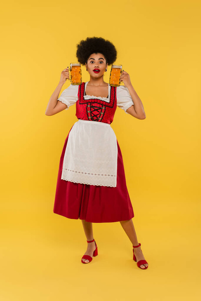 impresionada camarera afroamericana en traje bavariano tradicional sosteniendo tazas de cerveza en amarillo - Foto, imagen
