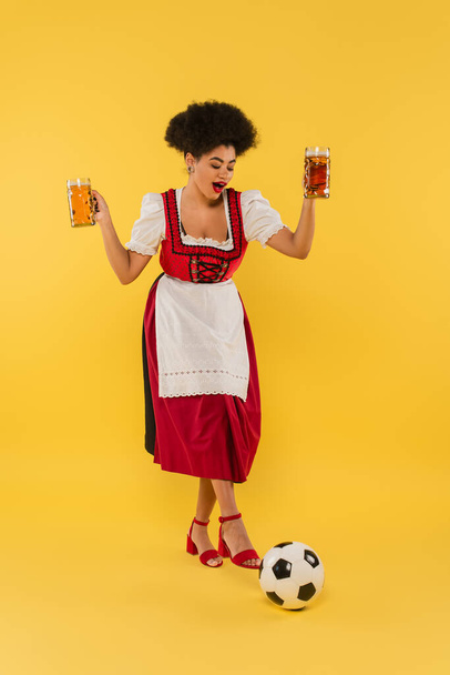 radosna afrykańska bawarska kelnerka w dirndl trzymająca kubki piwa w pobliżu piłki nożnej na żółtym - Zdjęcie, obraz