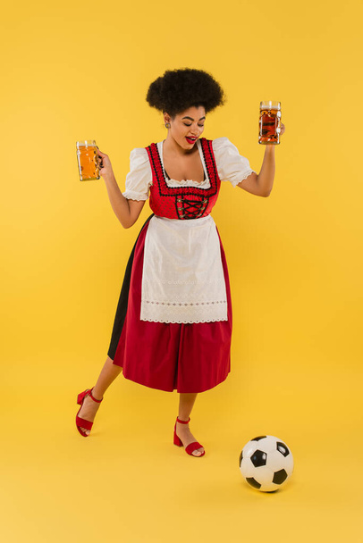 gioiosa cameriera afroamericana oktoberfest che tiene tazze di birra e gioca a calcio sul giallo - Foto, immagini