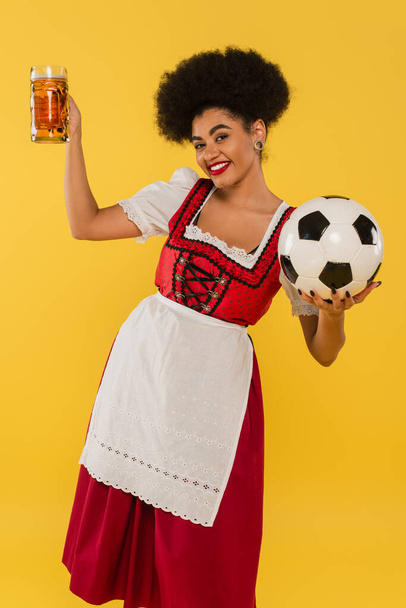 bezstarostná afroamerická servírka oktoberfestu v dirndl s pivním hrnkem a fotbalovým míčkem na žluté - Fotografie, Obrázek