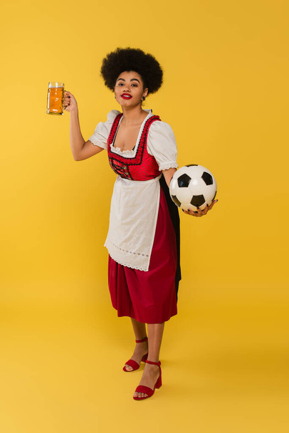 compiaciuta cameriera afroamericana in costume bavarese con boccale di birra e pallone da calcio su giallo - Foto, immagini