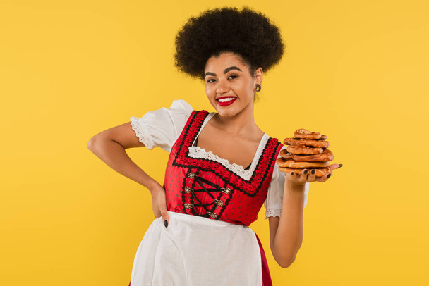 Afroameričanka oktoberfest servírka v tradičním kostýmu držící preclíky a usmívající se na žlutou - Fotografie, Obrázek